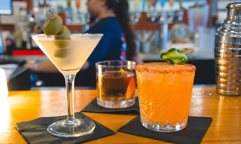 Three cocktails, a martini, an old fashioned, and an orange drink, on a wooden bar.