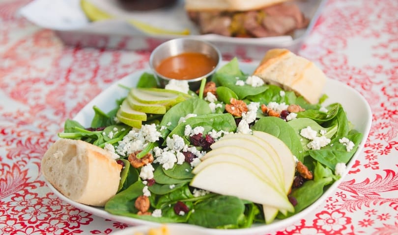 Spinach salad with sliced pears, blue cheese, cranberries, pecans, and dressing on a red patterned tablecloth.